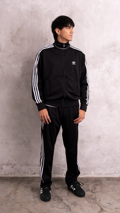 A person wearing a black Adidas tracksuit stands in a plain, gray-toned setting.