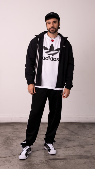 A man wearing an Adidas t-shirt, black jacket, and pants, standing in a plain room with a concrete floor.