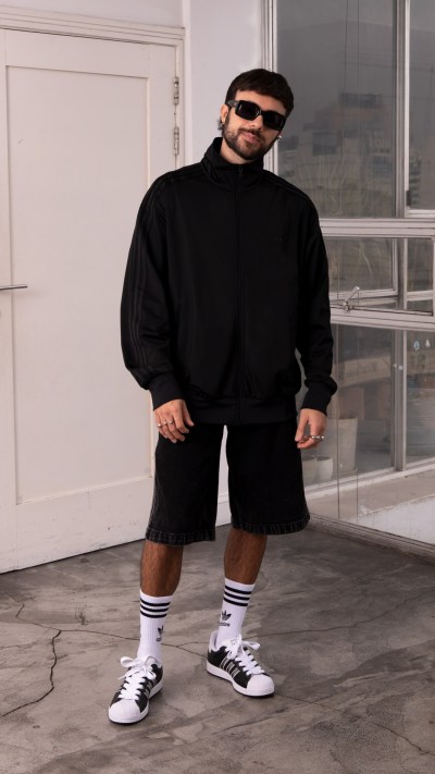 A person wearing a black sweater, shorts, and white sneakers stands in a room with a window in the background.