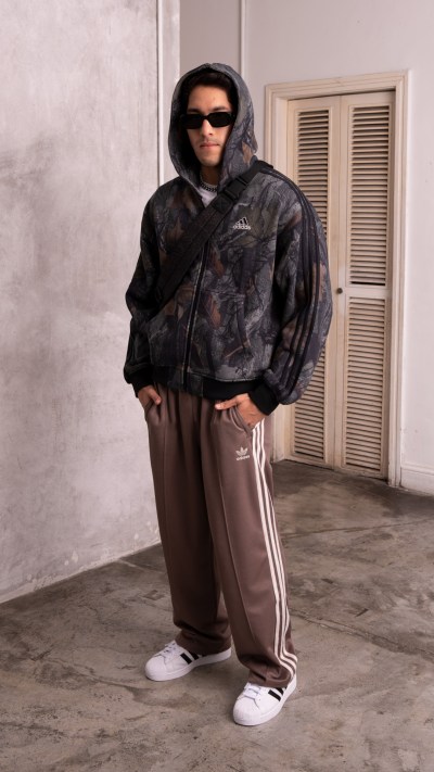 A person wearing a hooded camouflage jacket and sweatpants stands in a room with a concrete floor and a set of louvered doors in the background.
