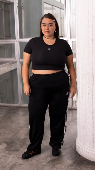 The image shows a person wearing a black crop top and black sweatpants, standing in a room with large windows and a concrete floor.