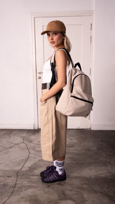 A young woman wearing a beige outfit, including a backpack, stands in a room with a plain white wall in the background.