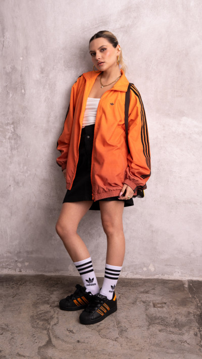 A person wearing an orange jacket, a white top, and black shorts stands in front of a plain gray wall.