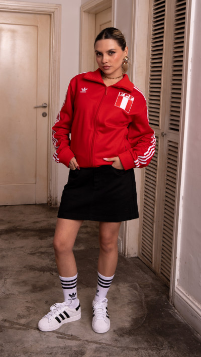 A person wearing a red jacket and black skirt stands in a room with a door and window visible in the background.