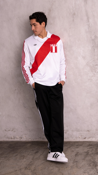 A young man wearing a white and red sports jersey stands against a plain gray wall, with black pants and white sneakers.