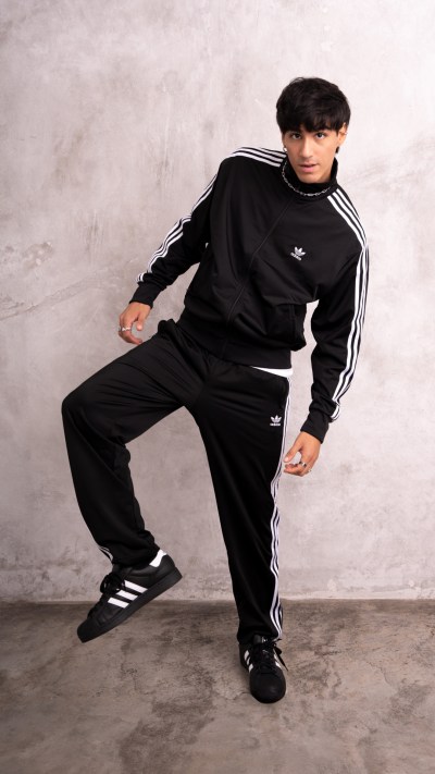 A young man wearing a black Adidas tracksuit stands against a plain, light-colored background, striking a casual pose.