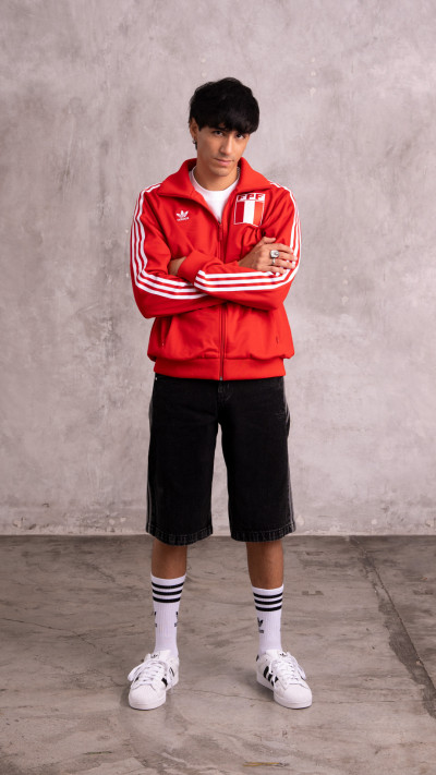 A young person wearing a red sports jacket and black shorts stands in front of a plain concrete wall.