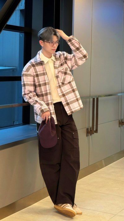 A young person wearing a plaid shirt, brown pants, and light-colored shoes stands in a room with glass walls and a tiled floor.