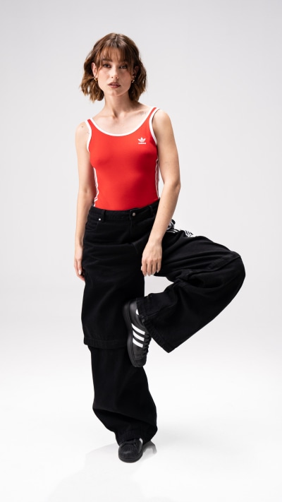 A young woman with curly hair wearing a red tank top and black pants stands against a plain white background, her expression conveying a sense of confidence and energy.