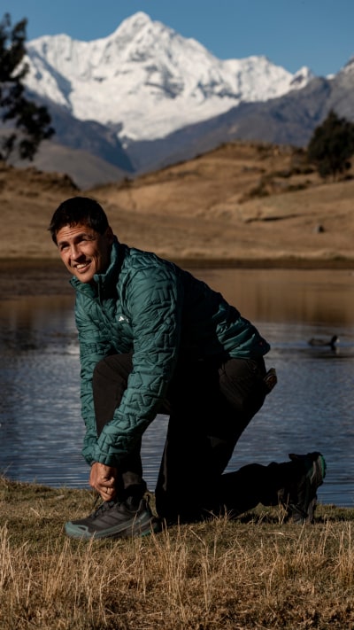 A person in a green jacket crouches in a grassy field, with snow-capped mountains visible in the background.