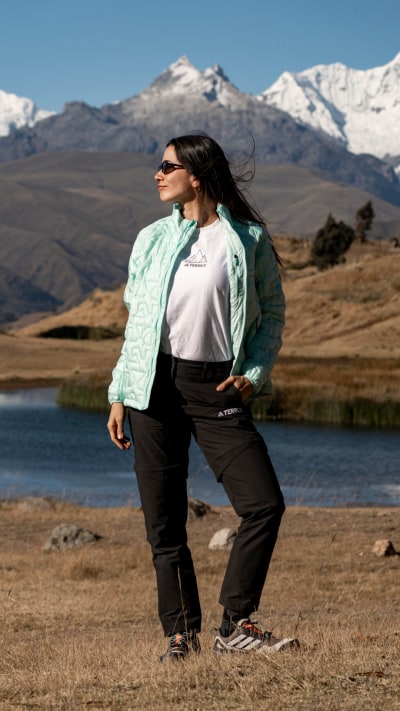 A person wearing a green jacket and sunglasses stands in a scenic landscape with snow-capped mountains in the background and a lake in the foreground.