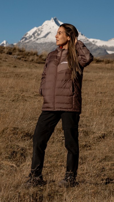 A woman in a brown puffer jacket stands in a grassy field, with a snow-capped mountain range visible in the background.