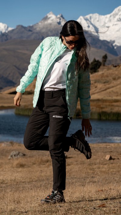 A person wearing a green jacket and black pants is crouching on a rocky shore, with snow-capped mountains visible in the background.