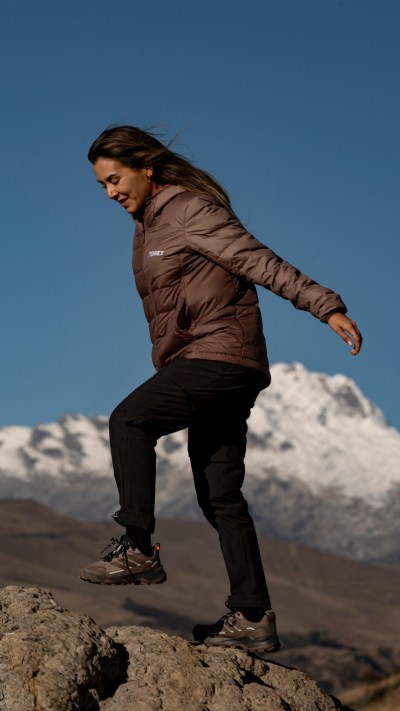 A person in a brown jacket and black pants is standing on a rocky outcrop, with snow-capped mountains visible in the background against a clear blue sky.