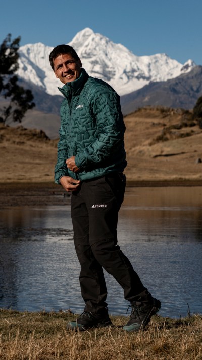 A person wearing a green jacket and black pants stands in a field with a lake and snow-capped mountains in the background.