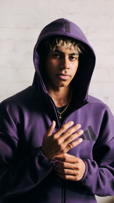 A young person wearing a purple hooded sweatshirt stands against a plain background, their hands clasped in front of them.