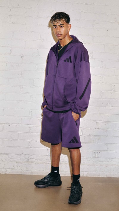 A young man wearing a purple tracksuit stands in front of a plain white wall.