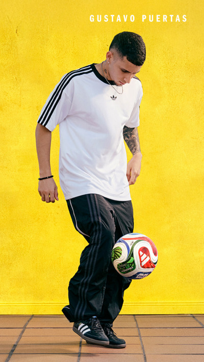 A young man wearing a white shirt and black pants is standing on a wooden floor, holding a soccer ball in front of a bright yellow background.