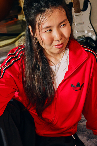 Mladá žena s dlhými tmavými vlasmi v žiarivo červenej bunde sedí pred gitarou a iným hudobným vybavením.