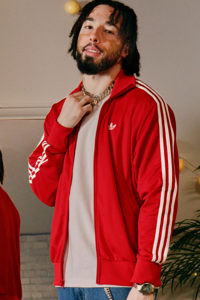 Ein Mann mit Dreadlocks in einer roten adidas Jacke steht in einem Raum mit einer weißen Wand und einer Pflanze im Hintergrund.