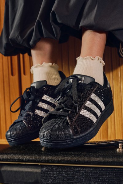 Čierno-biele tenisky adidas so šnúrkami položia na drevenom povrchu, pričom na pozadí vidno nohy človeka.