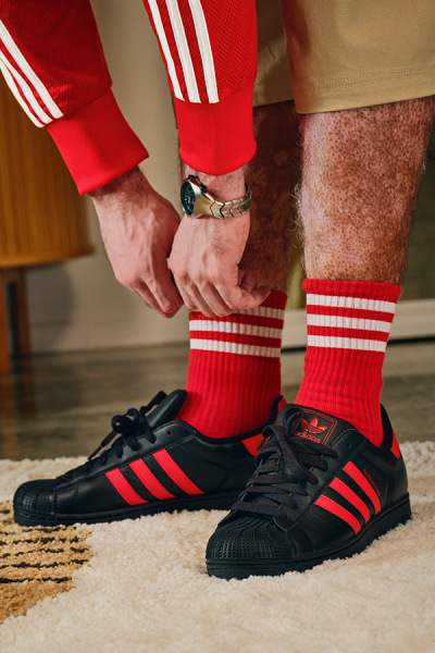 Das Bild zeigt eine Person im unteren Rumpfbereich, die rot-weiß gestreifte Socken, schwarz-rote Sneaker und rote sportliche Hosen trägt. Der Hintergrund scheint ein Teppichboden zu sein.
