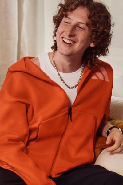 Usmievajúca sa osoba s kučeravými vlasmi v oranžovej bunde sedí v miestnosti na svetlom pozadí.