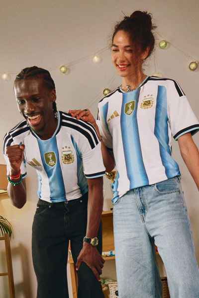 Dvaja ľudia, muž a žena, nosia futbalový dres argentínskeho národného tímu a s šťastným úsmevom sa usmievajú na tom, čo sa zdá byť domovom, keď na pozadí svietia šnúrky.