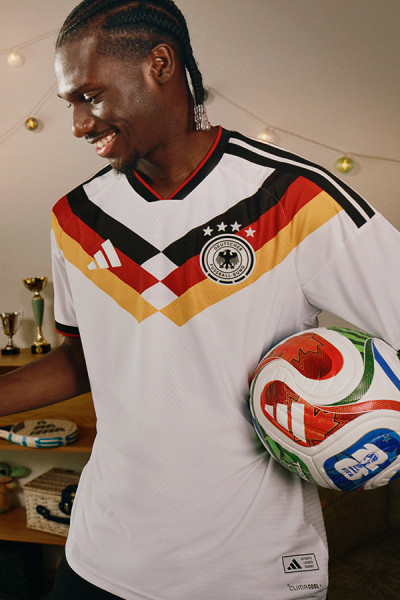 Das Bild zeigt einen Mann in einem weiß-rot-schwarzen Fußballtrikot mit dem Logo der deutschen Nationalelf, der einen Fußball vor einem Hintergrund mit verschiedenen Sporttrophäen und Fanartikeln hält.