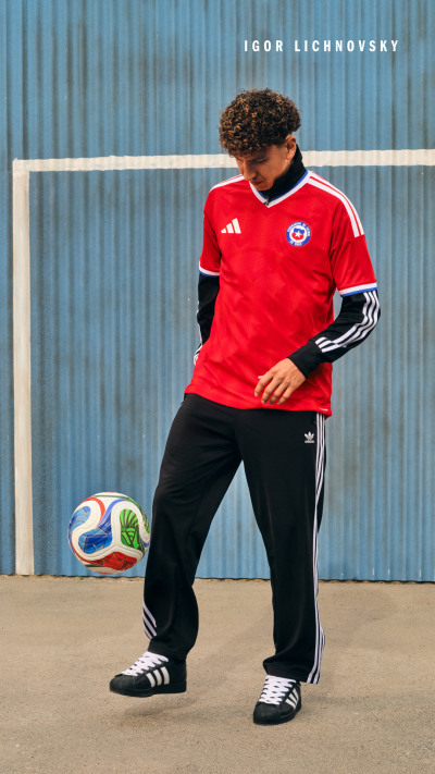 A person wearing a red soccer jersey and black pants stands in front of a blue metal wall, holding a soccer ball.