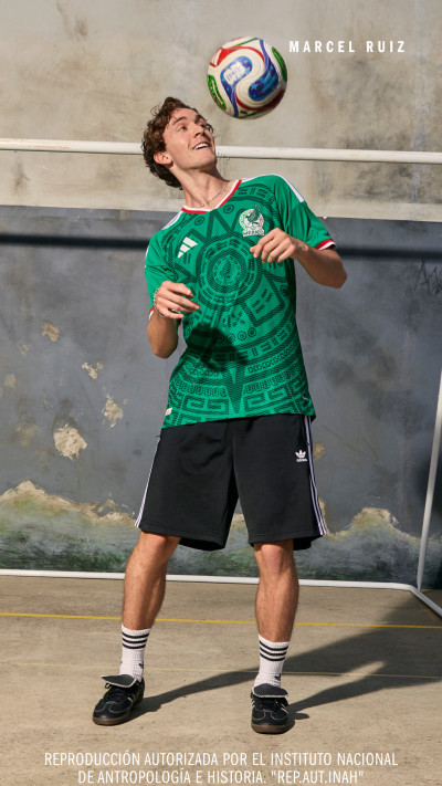 A young man in a green jersey with a soccer ball is standing in front of a mural depicting a scenic landscape.
