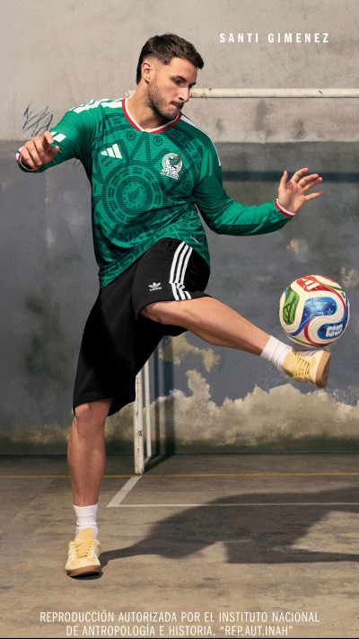 A soccer player in a green jersey is mid-kick, with a soccer ball in the air, against a wall with the name "Santi Gimenez" written on it.