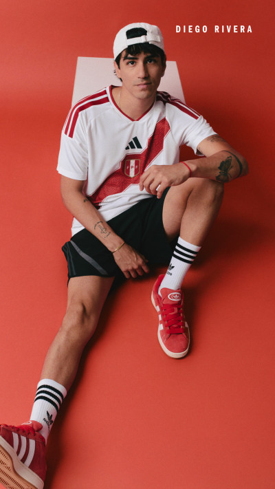 A young man wearing a white sports jersey, black shorts, and red sneakers is sitting on a bright red background.