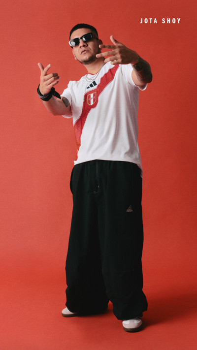 A person wearing a white t-shirt with a Peruvian flag, sunglasses, and loose black pants is gesturing with their hands against a bright red background.