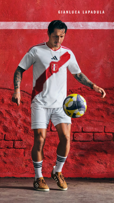 A young man in a white soccer jersey and shorts stands in front of a vibrant red brick wall, holding a soccer ball.