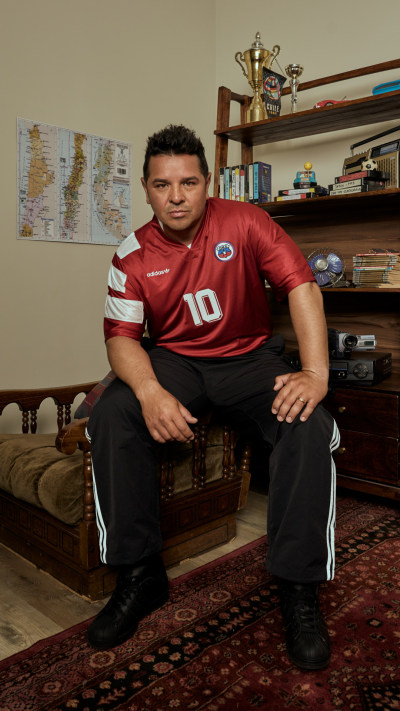 The image shows a man wearing a red sports jersey with the number 10 sitting on a couch in a room with bookshelves and a world map on the wall.