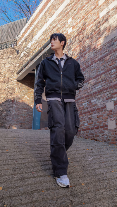 A person wearing a black jacket and pants stands in front of a brick building with a blue sky in the background.