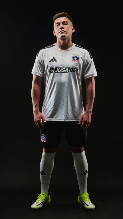 A muscular male athlete wearing a white soccer jersey with the "Juga" logo, standing in a dark background and wearing bright green cleats.