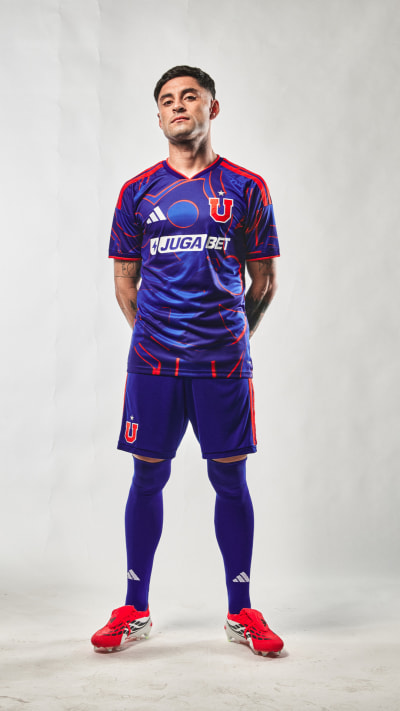 A young man wearing a purple and red soccer uniform stands confidently in front of a plain white background.
