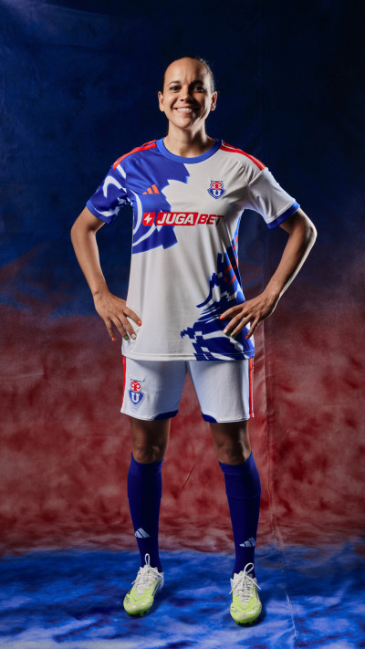 The image shows a smiling soccer player wearing a white and blue jersey, standing on a field with a red and blue background.