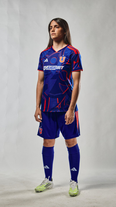 A young woman wearing a blue and red soccer jersey and shorts stands confidently against a plain background.