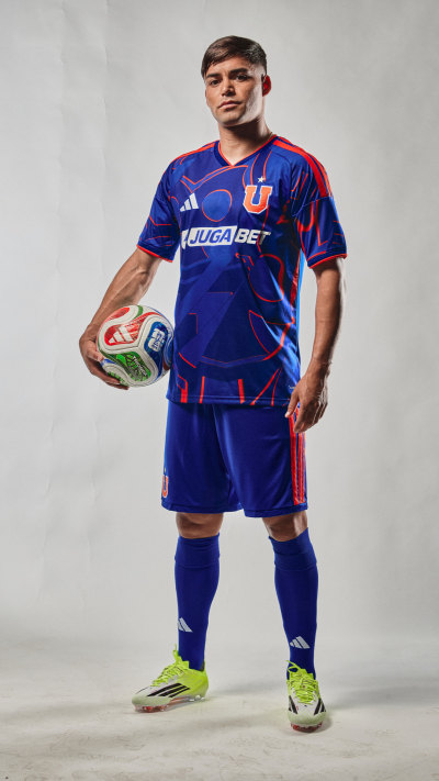 A man wearing a blue and red soccer jersey and shorts, holding a soccer ball, stands against a plain background.