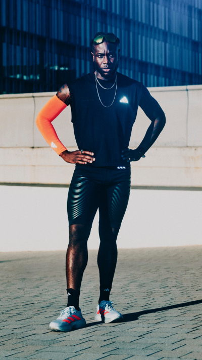 A person wearing a black outfit, including a turtleneck and leggings, stands on a wooden platform against a backdrop of blue metal panels.