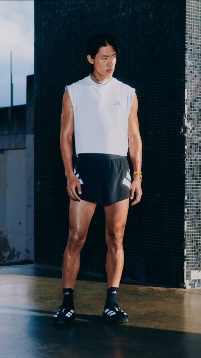 A muscular individual wearing a white sleeveless top and black shorts stands in front of a dark background, appearing to be in a fitness or training setting.