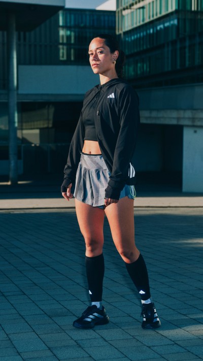 A young woman wearing a black crop top and gray shorts stands on a concrete surface, with a modern building in the background.