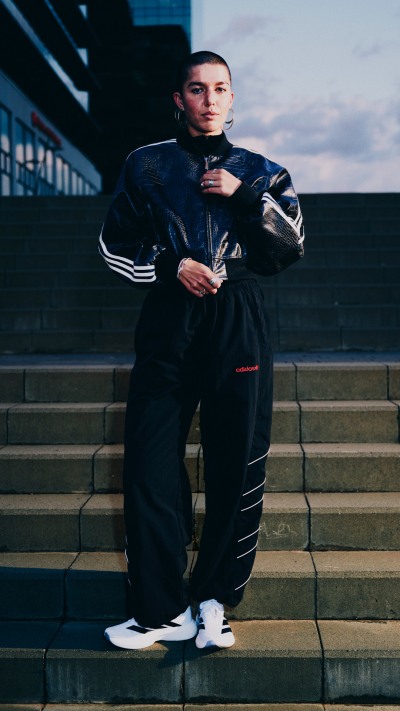 A person wearing a black tracksuit stands on a set of stairs, with a cloudy sky visible in the background.