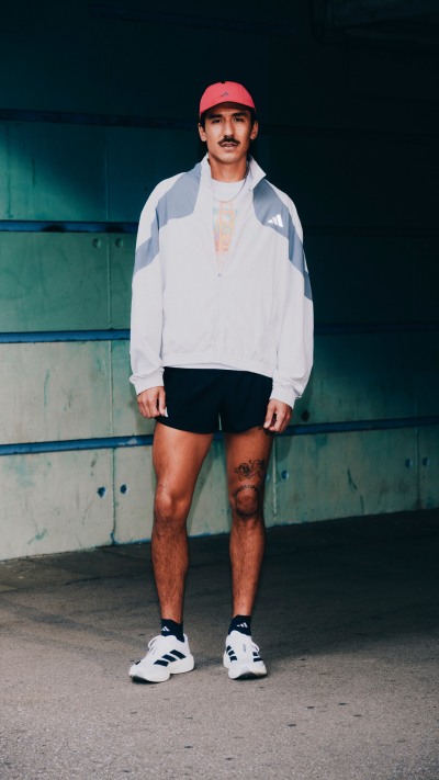 A person wearing a white sweatshirt, red cap, and sneakers stands in front of a green and blue wall.