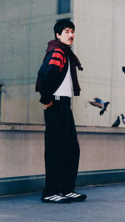 A person wearing a black and red striped jacket stands in front of a concrete wall, with pigeons visible in the background.