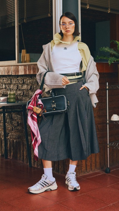 A young woman wearing a light-colored sweater and a dark skirt stands in front of a wooden structure, holding a skateboard and a backpack.