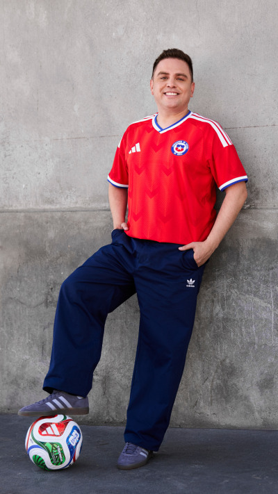 A smiling young man wearing a red sports jersey and navy blue pants is standing next to a soccer ball against a gray concrete wall.